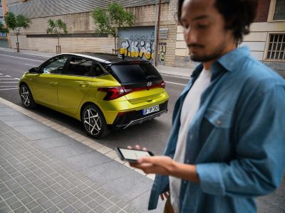Ein Mann überprüft sein Smartphone hinter dem Hyundai i20 in Gelb, der in einer Straße geparkt ist.