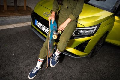 Eine Person, die ein Skateboard in der Hand hält und sich auf die Motorhaube eines Hyundai i20 stützt.